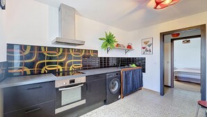 Cocina compacta con estufa, horno y lavadora, una planta, y una puerta que da a una zona de descanso.