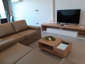 Una sala de estar moderna con un sofá, una mesa de centro con un arreglo floral, un televisor sobre un mueble de madera y una pared blanca con una ventana.