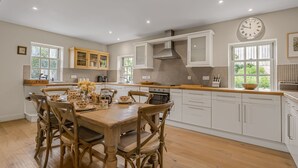 A spacious kitchen with white cabinetry, a wooden dining table, and a large wall clock. - Claytons Cottage -  Cotswold Family Retreat near Stow-on-the-Wold (Lower Oddington)