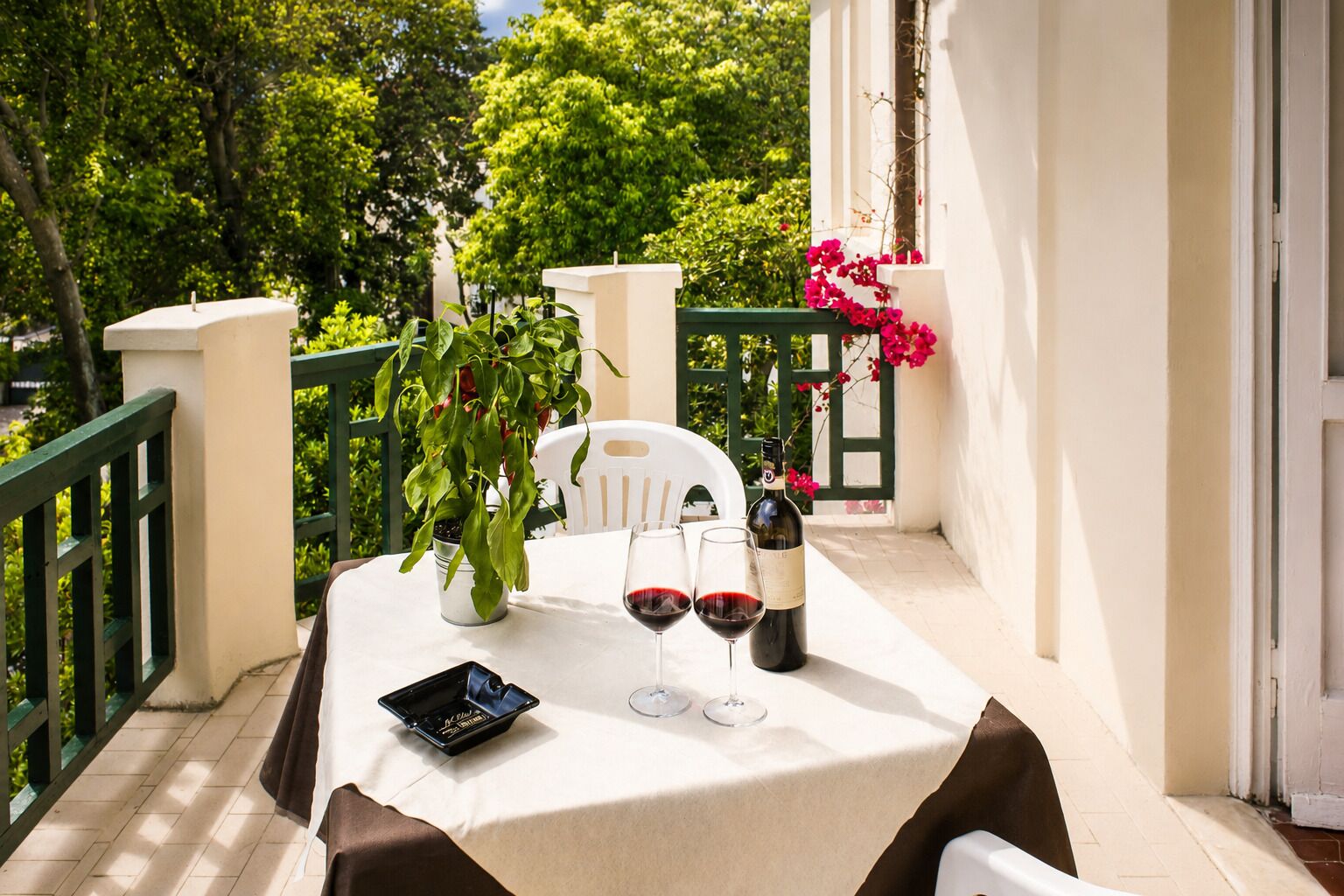 Una mesa al aire libre con una botella de vino, dos copas de vino, un teléfono inteligente y una planta en maceta en un balcón.