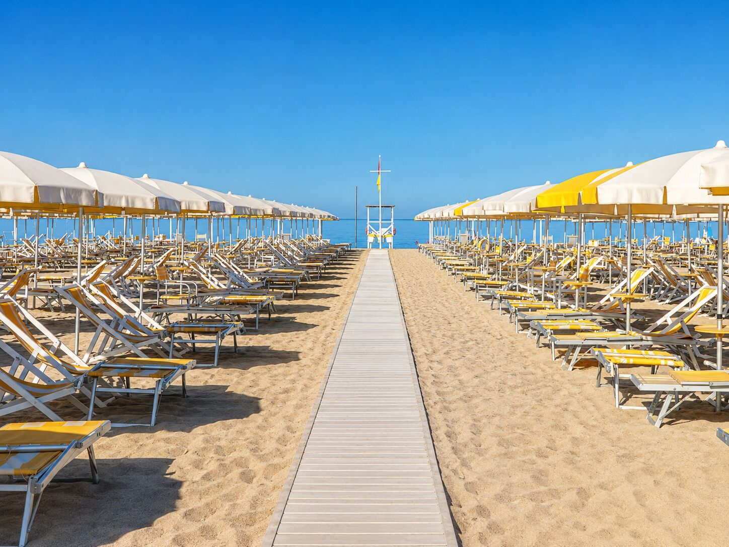 A beach with rows of sun loungers and umbrellas, a wooden walkway, and a lifeguard tower in the distance.