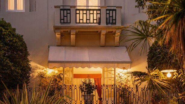 A two-story building with a balcony, a covered entrance, and a garden area with plants and seating. - Hotel Club i Pini- Residenza d'Epoca (Camaiore)