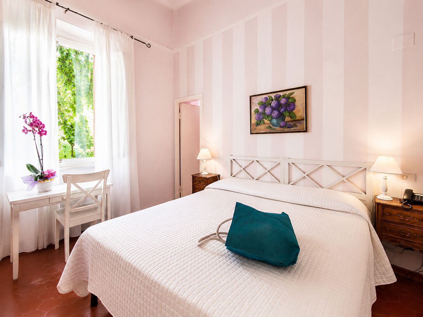 A hotel room with a bed, a desk, a chair, a window with curtains, a painting, and bedside lamps.