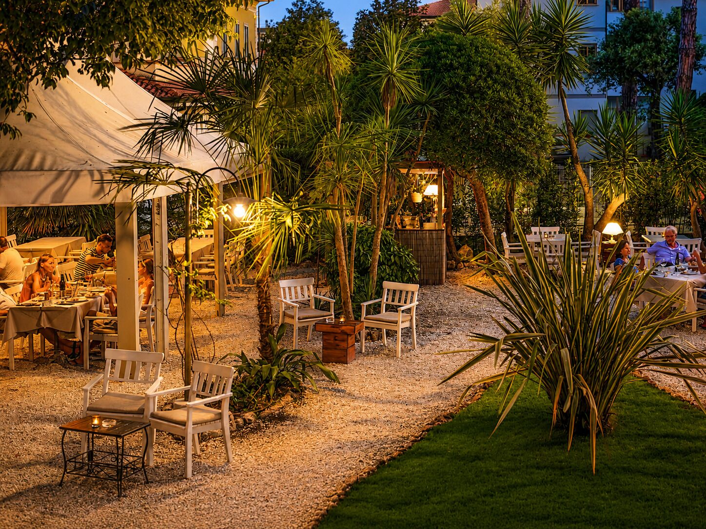 Un espace de restauration en plein air, avec des tables et des chaises, entouré de palmiers et bordé d’un chemin de gravier.