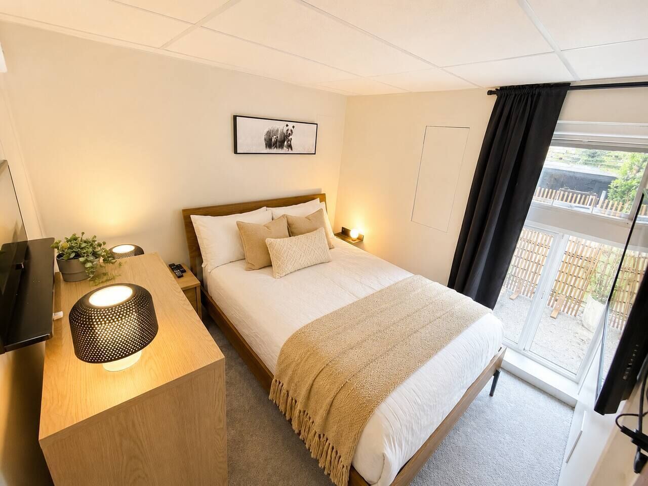 A bedroom with a bed, bedside table, lamp, and a view of the outside.