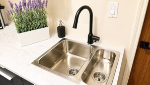 A kitchen with a double stainless steel sink, a black faucet, a soap dispenser, and a potted lavender plant. - Modern North Van Escape with 2BR | BBQ | Workspace (North Vancouver)