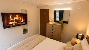 A bedroom with a bed, a dresser, a TV displaying a fireplace, a mirror, and a nightstand with a lamp. - Modern North Van Escape with 2BR | BBQ | Workspace (North Vancouver)
