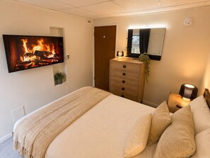 A bedroom with a bed, a dresser, a TV displaying a fireplace, a mirror, and a nightstand with a lamp. - Modern North Van Escape with 2BR | BBQ | Workspace (North Vancouver)