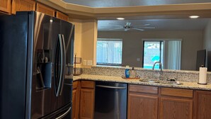 Una cocina con refrigerador de acero inoxidable, armarios de madera, encimeras de granito, lavavajillas, ventilador de techo y una ventana con persianas.