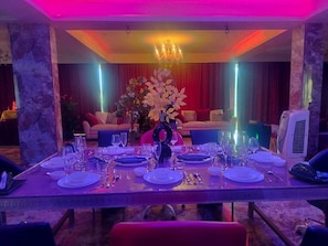 A dining area with a long table set for a meal, a chandelier, and a red curtain backdrop.