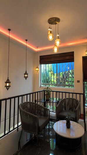 A room with a balcony, wicker chairs, a round table, hanging pendant lights, and a view of greenery through a window.