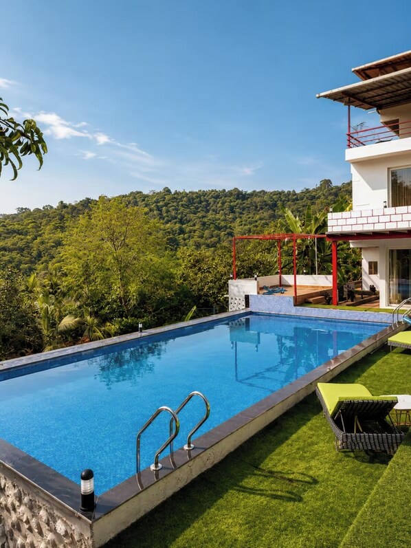 A swimming pool with a red structure, surrounded by lush greenery and a hill in the background.