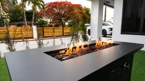 Un foyer extérieur moderne au design épuré et de couleur noire, entouré d’une pelouse bien entretenue et d’une clôture en bois.