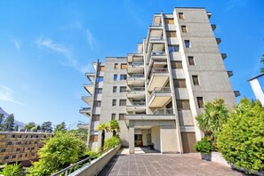 Ein mehrgeschossiges Gebäude mit Balkonen, einer gepflasterten Eingangszone und umliegender Bepflanzung.