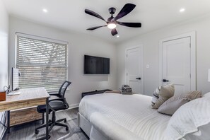 Une chambre à coucher comprenant un lit, un bureau, une chaise, un ventilateur de plafond et un téléviseur fixé au mur.