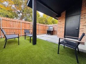 An outdoor patio area with artificial grass, two black lounge chairs, a barbecue grill, and a brick wall. - Stylish Dallas Escape - 3 Min to Love Field (Dallas)