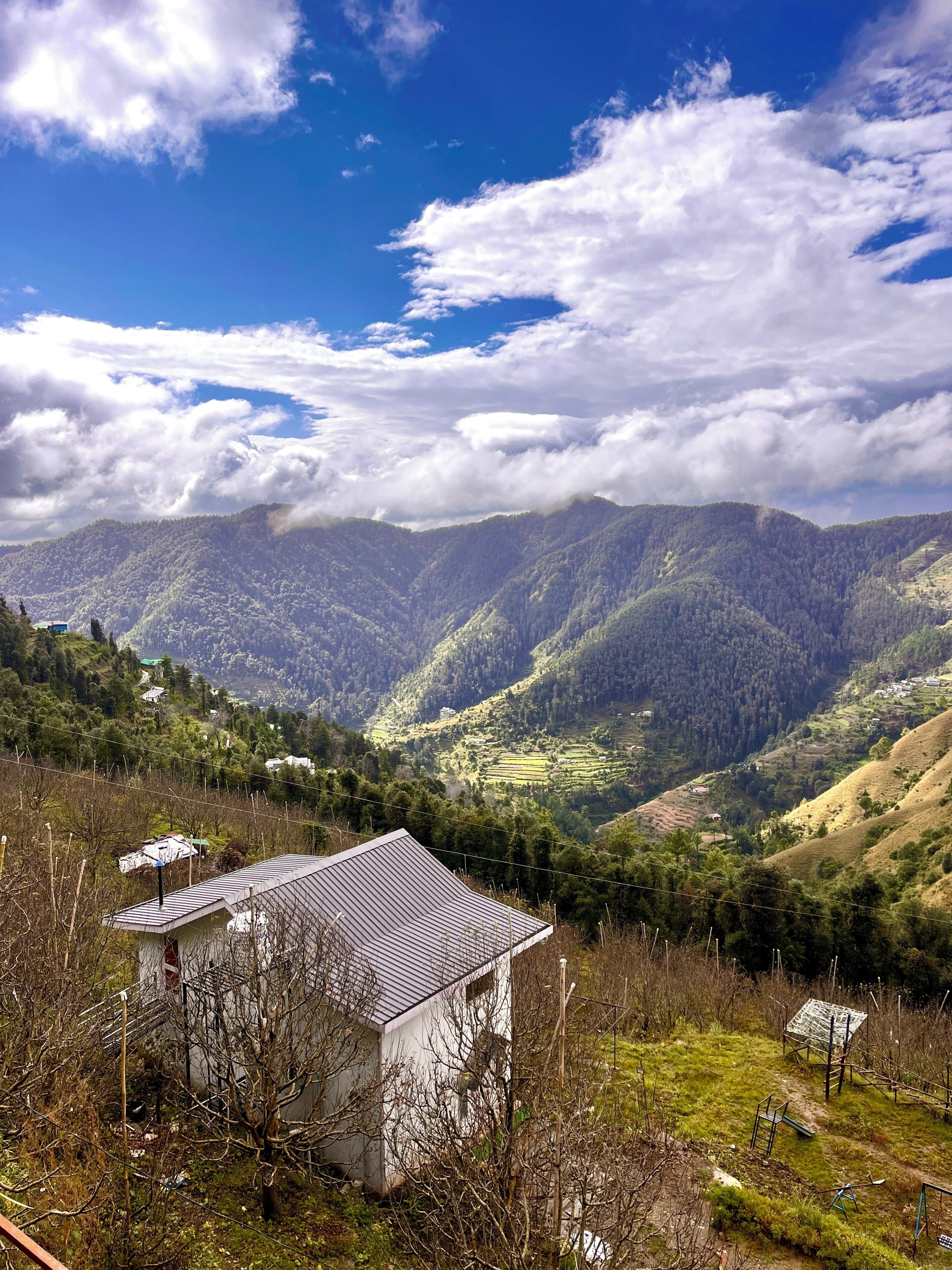 Paisaje montañoso con un pequeño edificio en primer plano, rodeado de vegetación exuberante y colinas distantes.