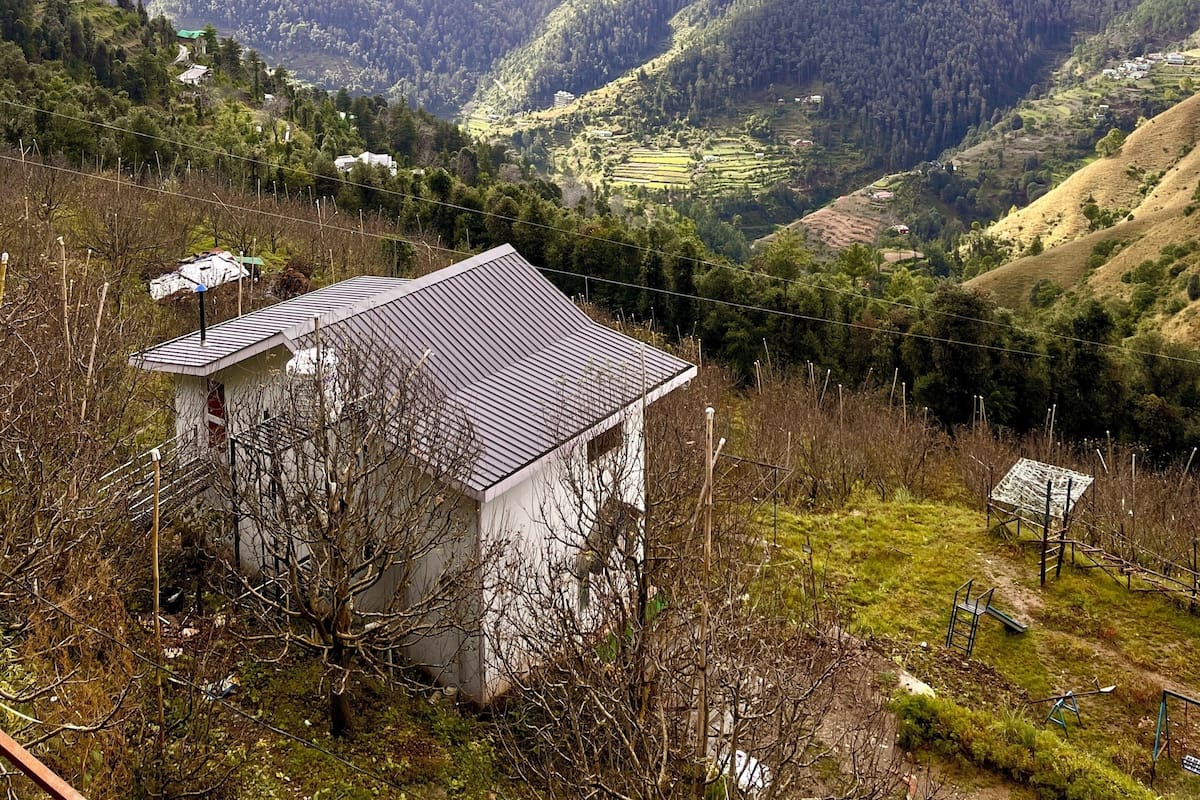 Paisaje montañoso con un pequeño edificio en primer plano, rodeado de vegetación exuberante y colinas distantes.