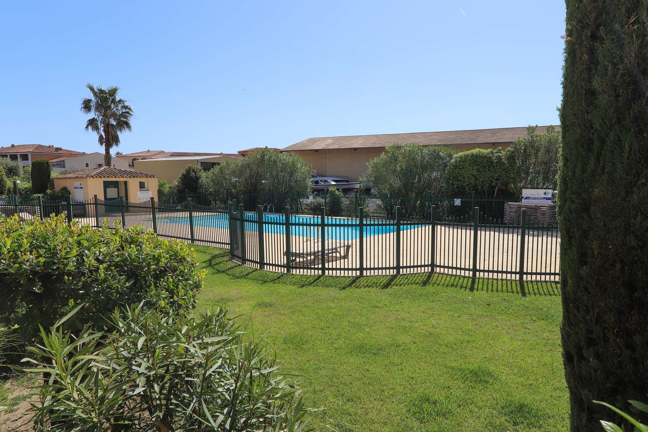 A fenced swimming pool area with a clear sky, surrounded by greenery and buildings.