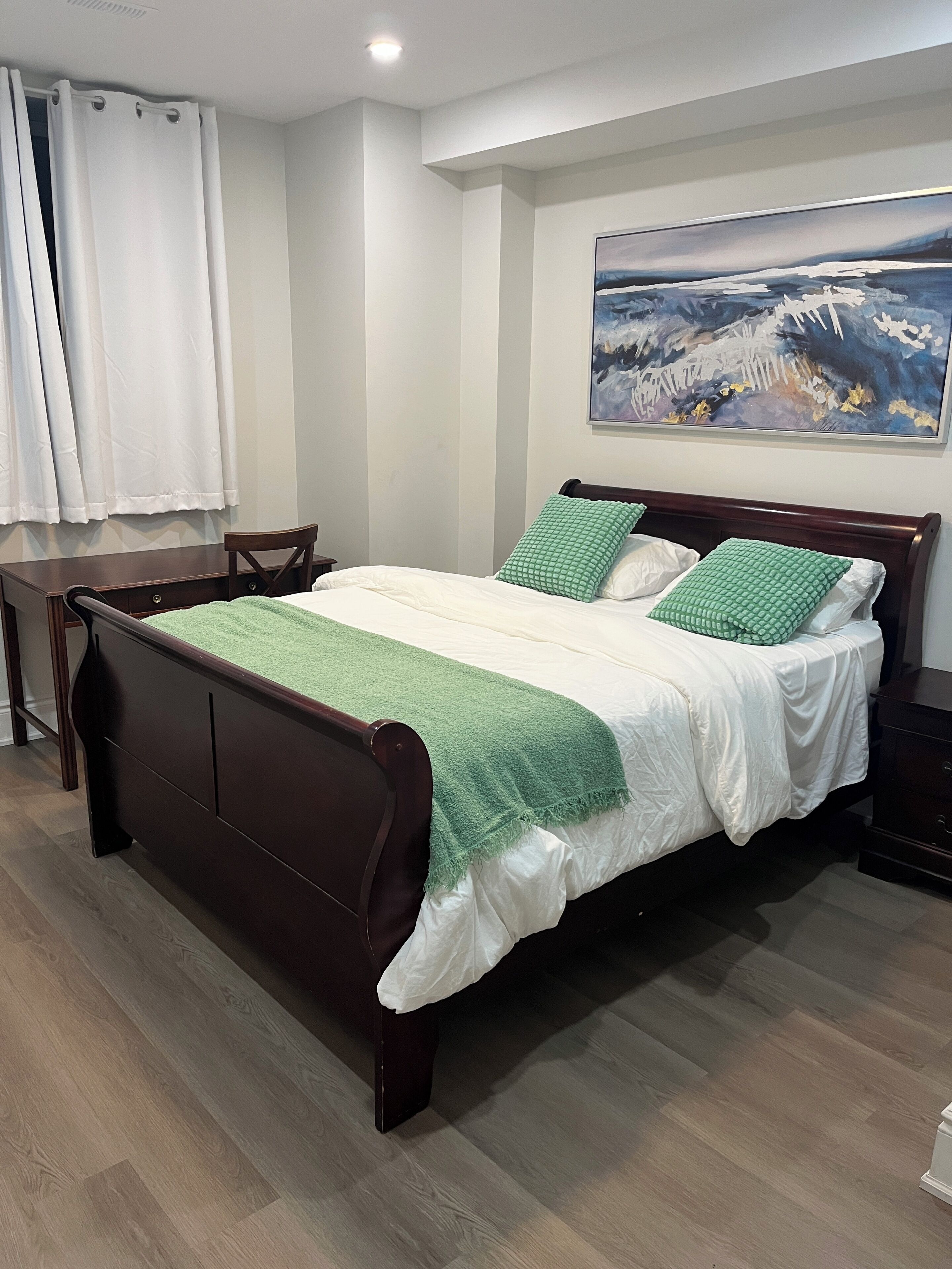 A bedroom with a wooden bed, a green and white bedspread, a painting on the wall, and a wooden floor.