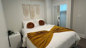 Ein Schlafzimmer mit einem Bett, weißen Wänden, Wanddeko aus Makramee, einer braunen Tagesdecke und einem Nachttisch mit einem Telefon.