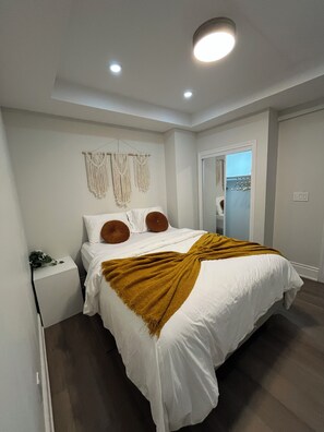 Ein Schlafzimmer mit einem Bett, weißen Wänden, Wanddeko aus Makramee, einer braunen Tagesdecke und einem Nachttisch mit einem Telefon.