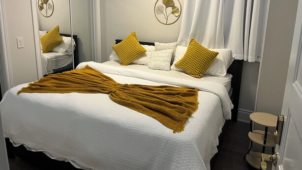 Ein ordentlich bezogenes Bett mit weißer Bettwäsche und einer senffarbenen Tagesdecke, ein Nachttisch mit Tablett sowie Wandbilder.