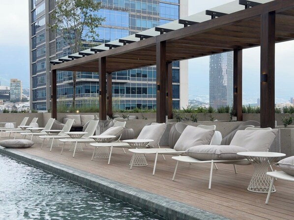 A rooftop pool area with lounge chairs and tables, a pergola structure, and a city skyline in the background.