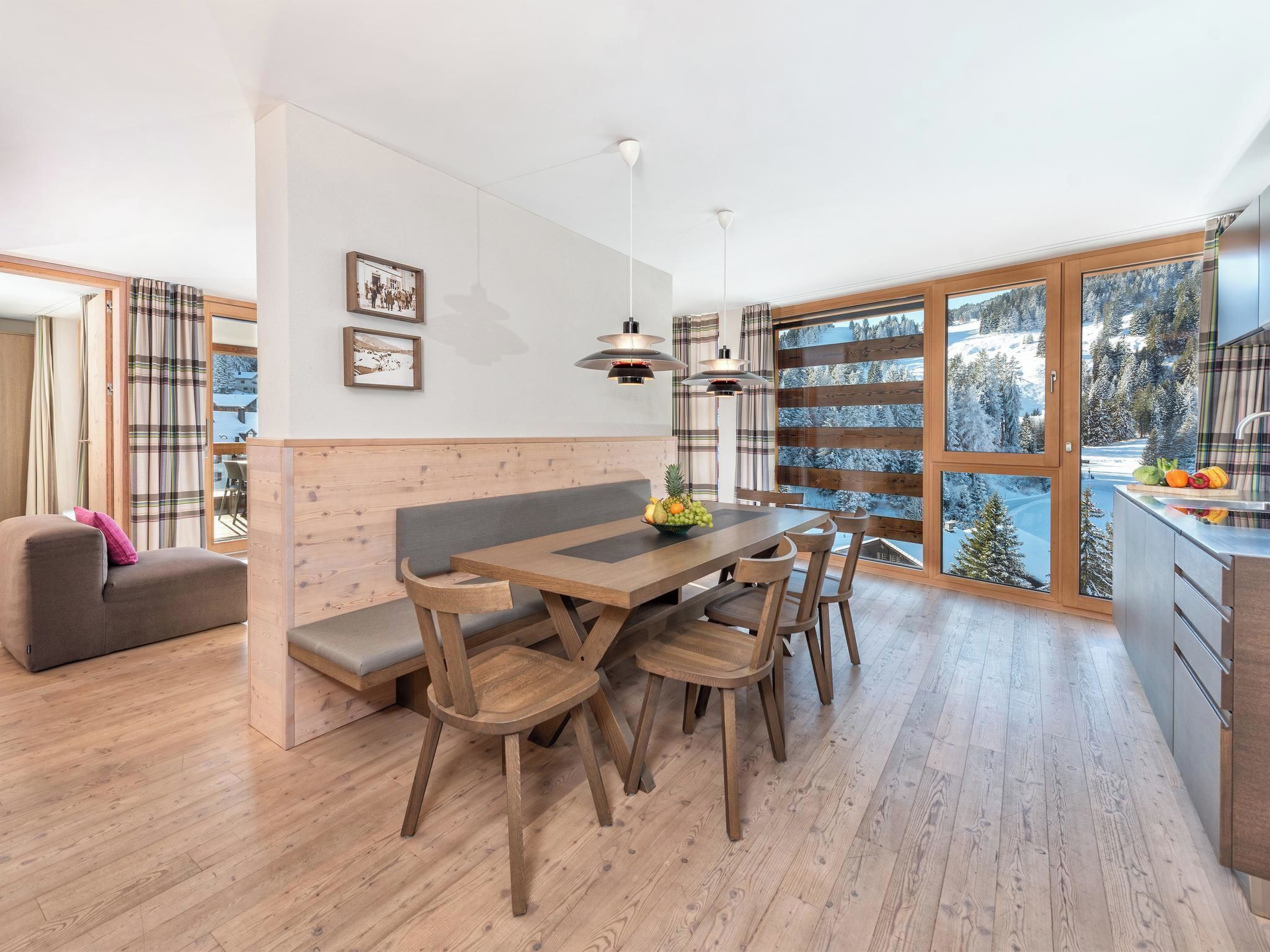Une cuisine moderne avec des meubles en bois, un coin repas doté d’une table et de chaises en bois, et de grandes fenêtres offrant une vue sur un paysage enneigé.