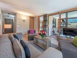 A modern living room with a sofa, armchair, and a coffee table. There is a TV and a window with a view of the outdoors.