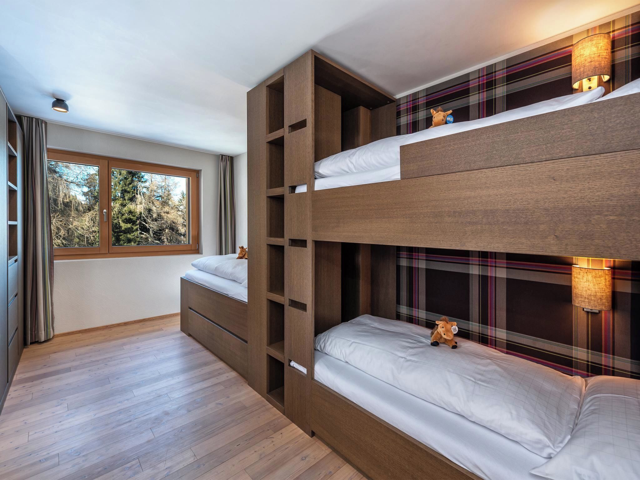 Um quarto com beliche, janela com vista para árvores e parede com padrão xadrez.