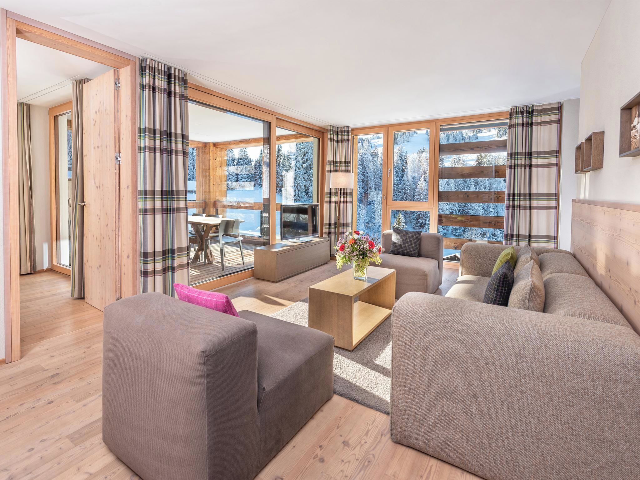 A spacious living room with a sofa set, a coffee table, and a TV. There is a large window with a view of snow-covered trees.