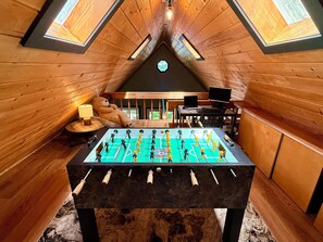 Ein Dachzimmer mit Holzwänden, einem Kicker, einem großen Teddybären und einem Schreibtisch mit einem Laptop.