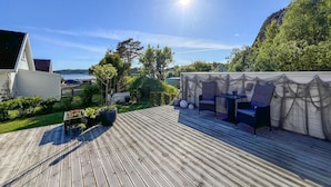 Eine Holzterrasse mit Gartenmöbeln, ein weißes Gebäude und Blick auf ein Gewässer.