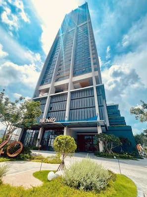 Un edificio moderno de gran altura con fachada de vidrio, una entrada destacada y un área ajardinada con vegetación al frente.