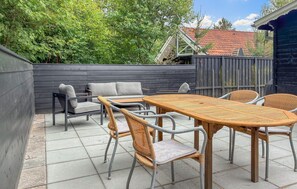 Eine Terrasse im Freien mit einem Holztisch und Korbstühlen, umgeben von einer niedrigen Mauer und Bäumen.