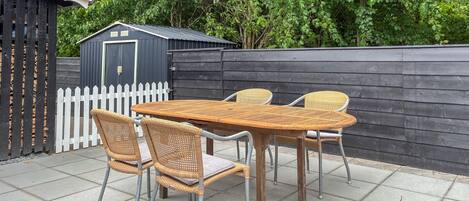 En terrasse med et træbord og fletstole, et hvidt stakittancehegn og en mørk trævæg.