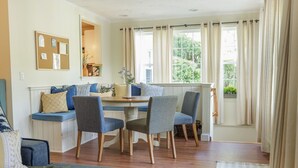 Una habitación acogedora con una mesa de madera, cuatro sillas, un banco con cojines, un sofá y una ventana con cortinas.