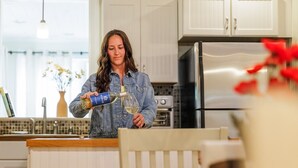 Una persona sirve una bebida en un vaso en una cocina con armarios blancos, un refrigerador y una ventana con vistas.
