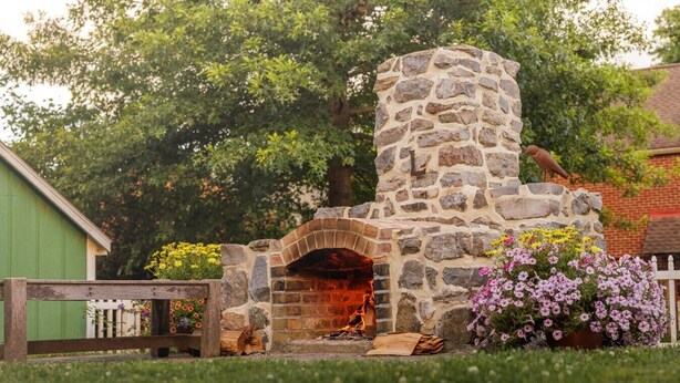 Un horno de pizza de piedra con arco de ladrillo, rodeado por una valla de madera, flores y un edificio verde al fondo.