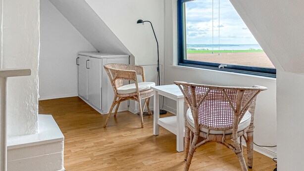 Une petite pièce de style « loft », aux murs blancs, dotée d’un plancher en bois, d’un bureau blanc, de deux chaises en osier, d’une armoire blanche et d’une fenêtre donnant sur un champ.