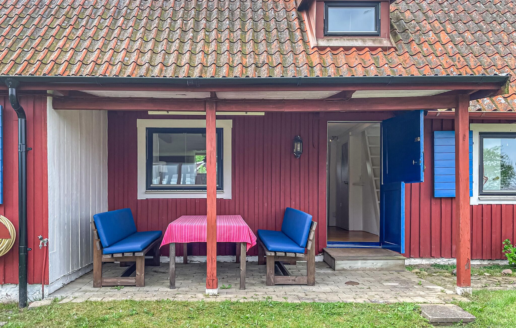 Un bâtiment rouge avec un toit en tuiles, un patio couvert avec deux chaises bleues et une table, et une porte donnant sur un espace intérieur.