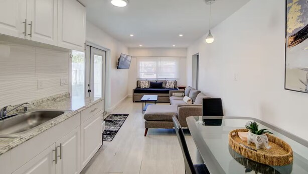 A modern kitchen with white cabinets, a granite countertop, and a dining area with a glass table and chairs. - Cozy 1-bedroom house in pleasant Pompano Beach with AC (Pompano Beach)