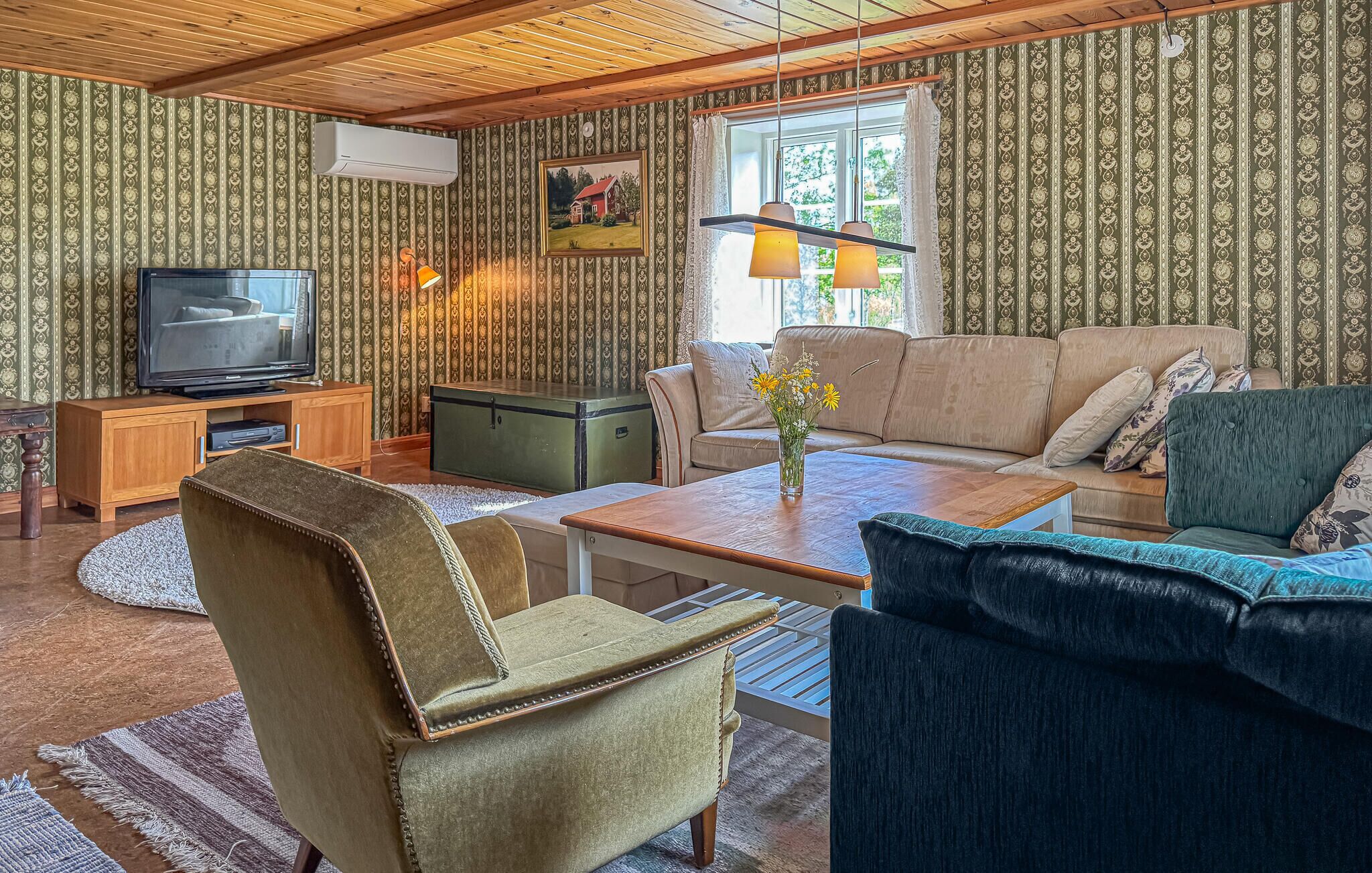 Ein gemütliches Wohnzimmer mit Fernseher, Sofa und Sessel, mit gemustertem Wandpapier und Holzbalkendecke.
