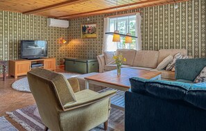 A cozy living room with a television, sofa, and armchair, featuring patterned wallpaper and wooden ceiling beams. - Gorgeous home in Aneby with WiFi (Aneby)