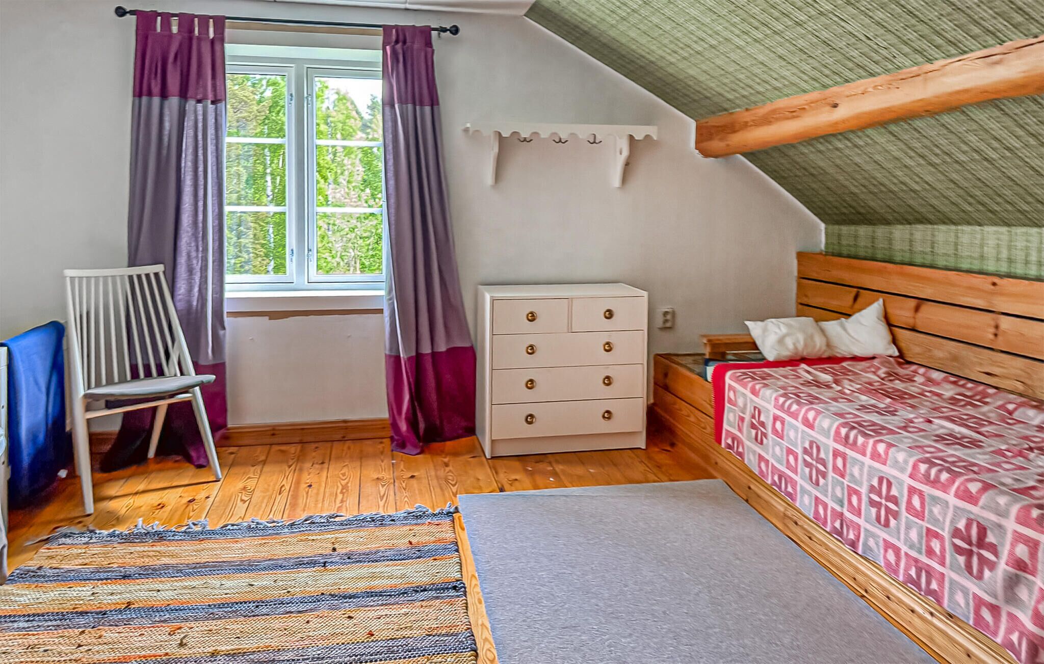 A bedroom with a wooden bed, a dresser, a chair, a window with curtains, a rug, and a sloped ceiling.
