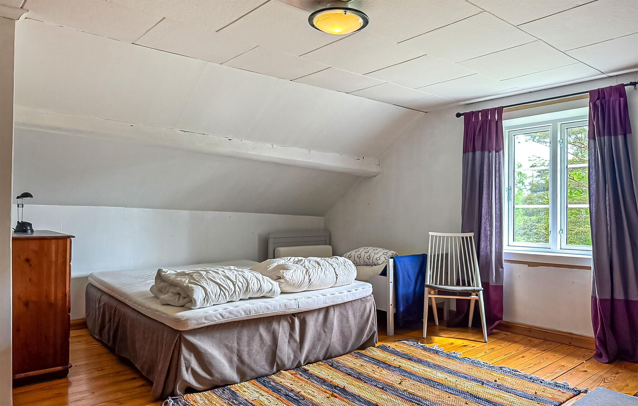 Un dormitorio con cama, escritorio, silla, ventana con cortinas e iluminación en el techo.