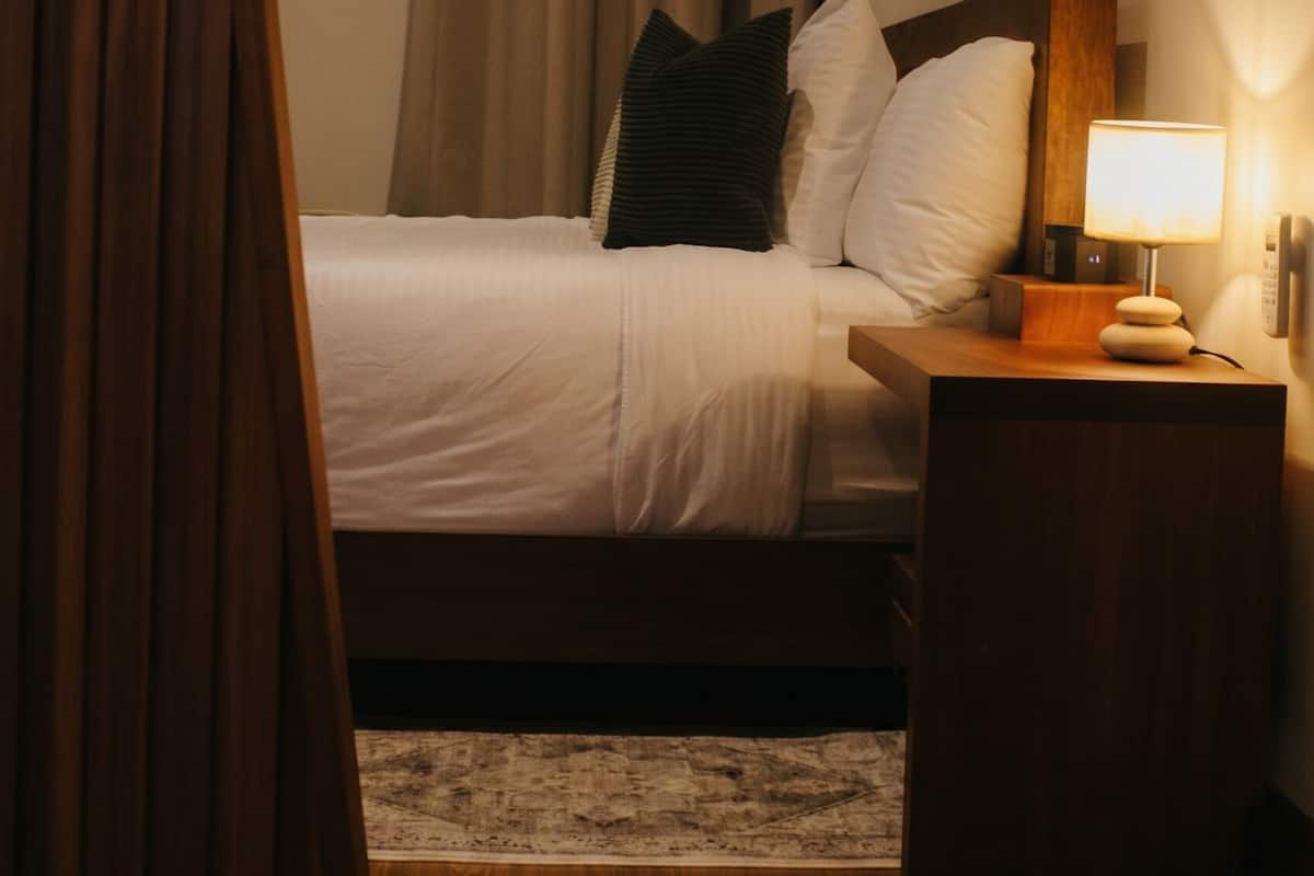 A hotel room with a bed, wooden nightstand, lamp, curtains, and a wooden floor.
