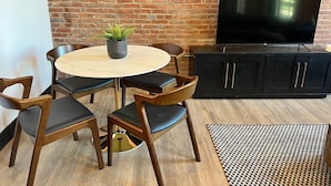 Een kamer met een bakstenen muur, een ronde tafel met stoelen, een zwarte kast en een televisie.