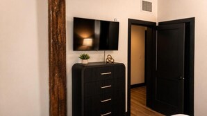 Een slaapkamer met een bed, een dressoir, een televisie aan de muur, en een deur naar een andere kamer.